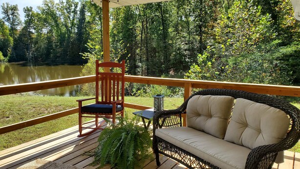 A wooden deck with a wicker sofa and a wooden chair, overlooking a lake and surrounded by trees.