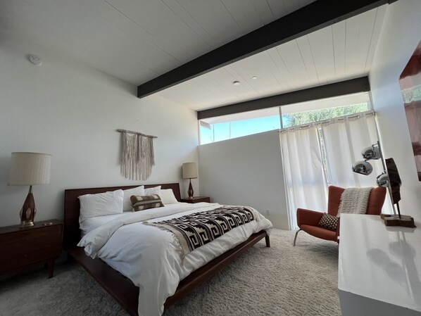 Quarto com uma cama grande, duas mesas de cabeceira, uma cadeira, uma janela com cortinas e uma luminária na parede.