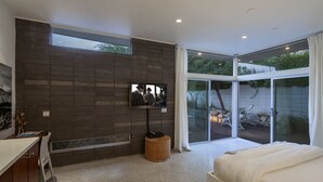 A modern bedroom with a large bed, a TV mounted on a brick wall, and a sliding glass door leading to an outdoor area. - The Butterfly 4 Bedroom Home, Ideal Downtown Historic Tennis Club location (Palm Springs)