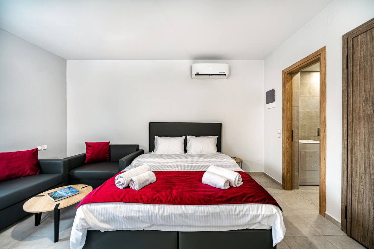 A modern hotel room with a bed, two red pillows, a red throw blanket, a small wooden table with a magazine, and a black sofa.