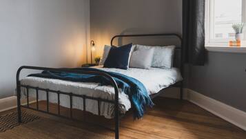 A single bed with a metal frame, white bedding, and a blue throw blanket. There is a bedside table with a lamp and a small potted plant. The room has wooden flooring and a window with a view of the outside.