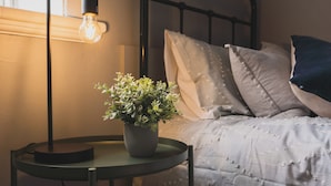 Un lit bien fait, recouvert d’une couvre-lit à motifs gris et blancs, une plante en pot posée sur une petite table ronde à côté, et une lampe de sol dont l’ampoule est visible.