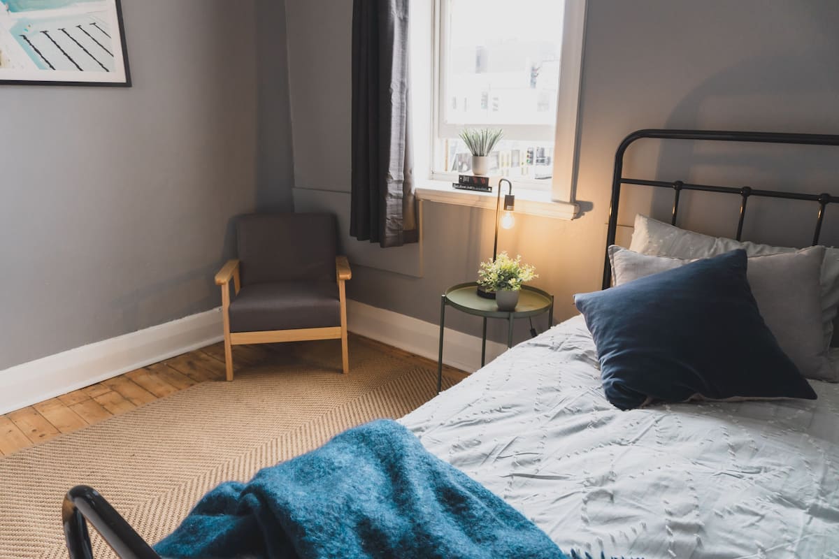 A bedroom with a bed, a chair, a small table with a plant, and a window with curtains.