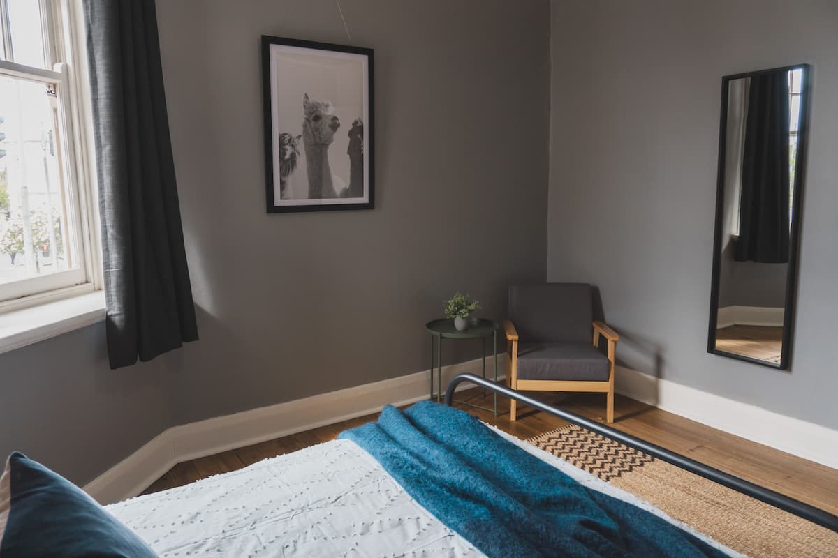 A bedroom with a bed, a chair, a small table with a plant, a framed picture on the wall, and a large mirror.