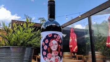 Una botella de vino con etiqueta floral, sobre una mesa al aire libre junto a una planta en maceta y luces colgantes en el fondo.