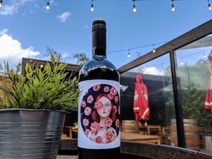 A bottle of wine with a floral label, placed on a table outdoors with a potted plant and string lights in the background. - Royal Derby Hotel (Fitzroy)