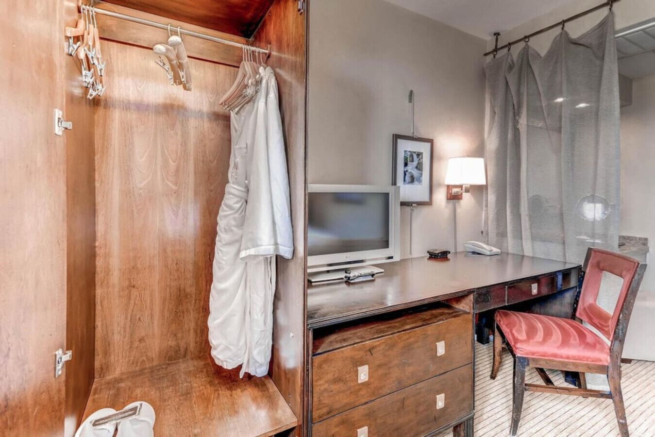 A hotel room with a wooden wardrobe, a desk with a television, a chair, and a bed.