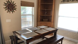 Een eethoek met een houten tafel en banken, een zondecoratie aan de muur, een raam met uitzicht op een balkon, en een houten kast met planken.