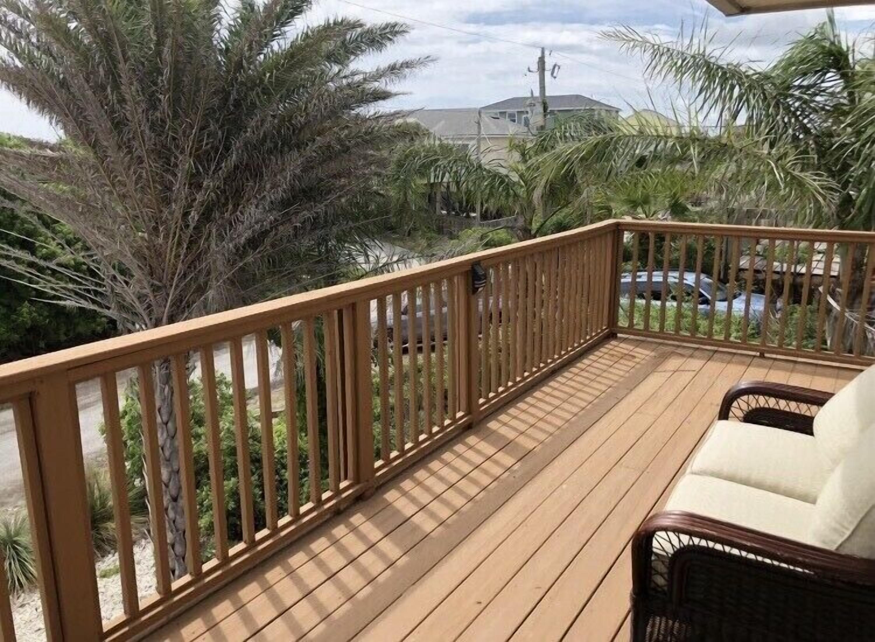 A wooden deck with a railing, a wicker chair, and a view of palm trees and a building in the background.