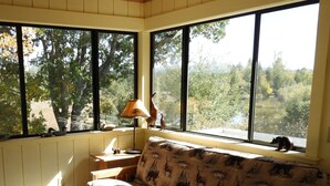 A cozy room with a wooden bench, a sofa with animal prints, a lamp, and large windows offering a view of trees and a lake. -  Cassel, Ca. on Hat Creek. Close/Baum Lake, Burney, Mt. Shasta/Lassen 3 nite min (Cassel)