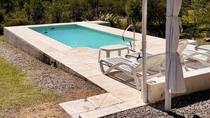 A rectangular swimming pool with a tiled deck, surrounded by gravel and grass. There are white outdoor chairs and a covered seating area with a pergola. The background features a clear blue sky and a hilly landscape. - Arroyo Suite House (Nono)