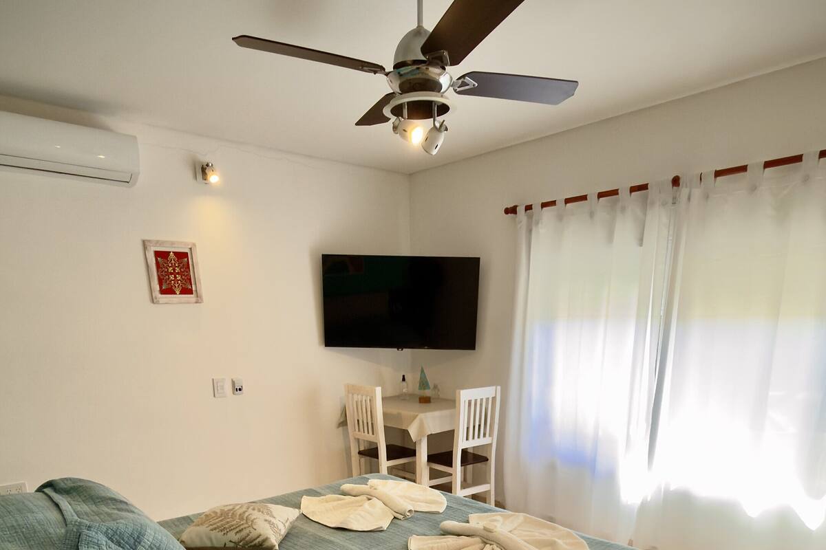 Un dormitorio con cama, ventilador de techo, televisor, mesa con sillas y ventana con cortinas.