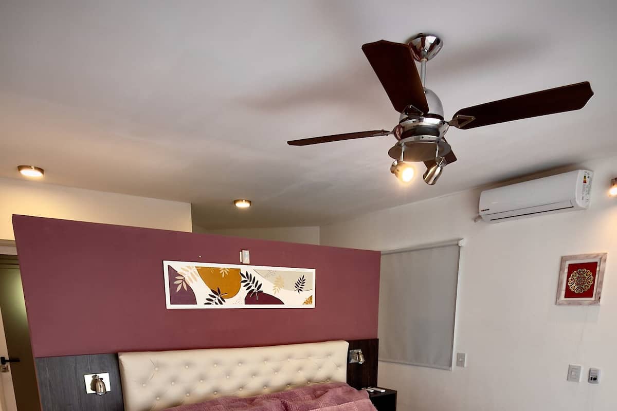 Un dormitorio con cama, ventilador de techo, mesita de noche, aire acondicionado de pared y un adorno en la pared.