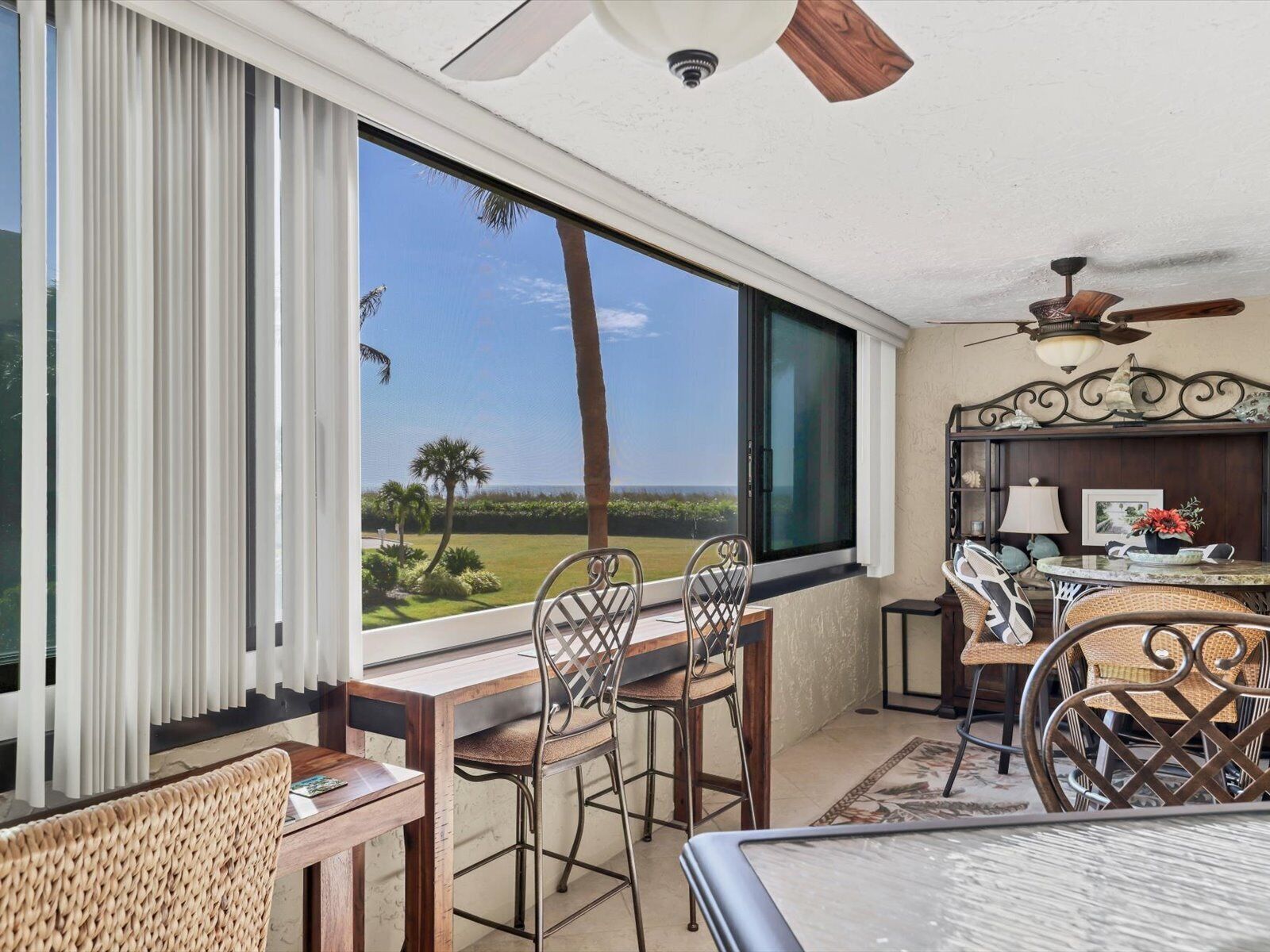 A room with a large window offering a view of the outdoors, a ceiling fan, and a dining area with wicker chairs and a table.