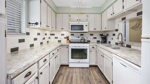 Una cucina con mobili bianchi, un forno a microonde, un forno, un lavello e una finestra con persiane.