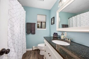 Une salle de bain avec des toilettes blanches, un lavabo doté d’un plan de travail foncé, un miroir et un rideau de douche à motifs géométriques.