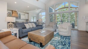 A spacious living room with a large sofa, a coffee table, and a patterned rug. There is a kitchen area in the background with white cabinets and a large window.