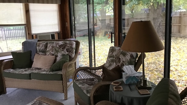 A cozy room with wicker furniture, a table with a lamp, and large windows offering a view of the outdoors.