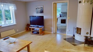 A living room with a flat-screen TV on a wooden stand, a dining table, and a kitchen visible through an open doorway. - Lovely cottage with large terrace by the sea and nature. (Bräkne-Hoby)