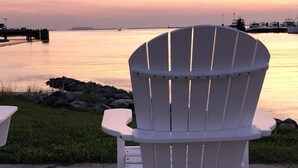 Una silla Adirondack blanca mirando a un cuerpo de agua tranquilo con un atardecer al fondo.