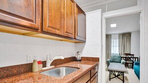 Una cocina con encimeras de granito, armarios de madera y un lavamanos de acero inoxidable. Se ve un área de comedor con una mesa y sillas a través de un vano abierto.