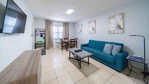 Una sala de estar con un sofá azul, una mesa de centro de madera, un televisor montado en la pared y un comedor con mesa y sillas.