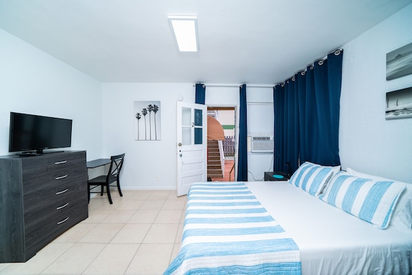 A bedroom with a bed, a desk, a chair, a dresser, a television, and a door leading to an outdoor area.