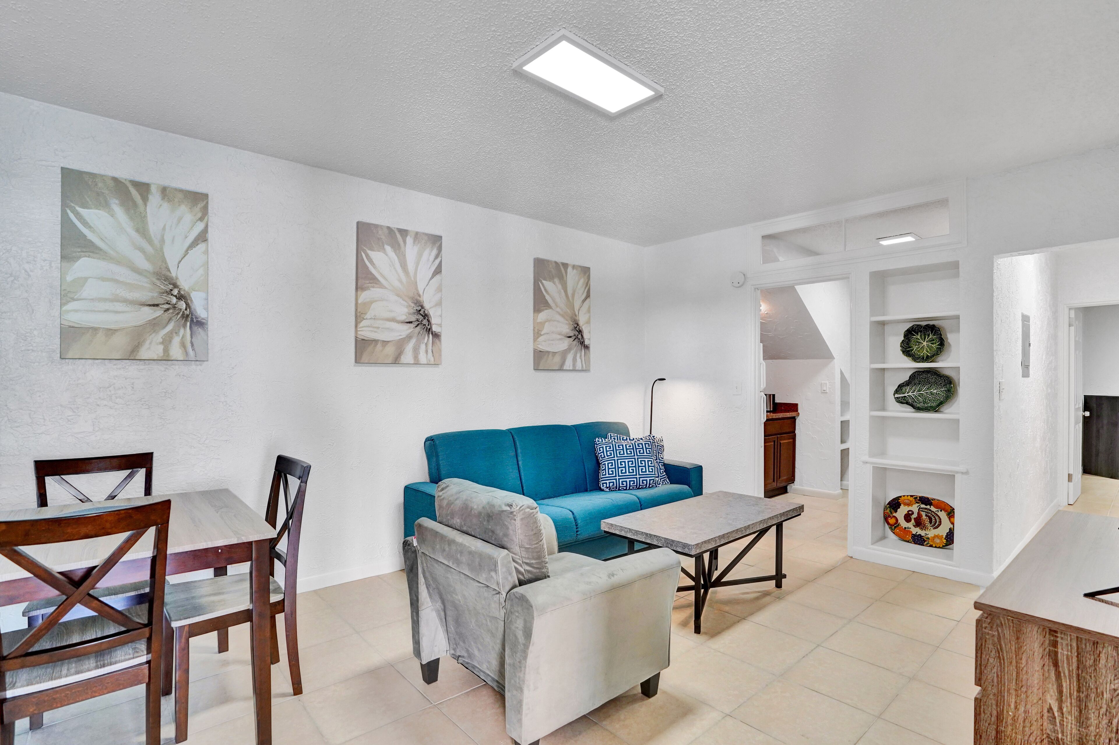 A modern living room with a blue sofa, a grey armchair, a wooden dining table, and a coffee table. There are abstract floral paintings on the walls and a white shelving unit with decorative items.