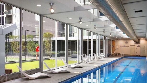 Una piscina interior con paredes de vidrio transparente, sillas de descanso y vistas a un espacio exterior verde.