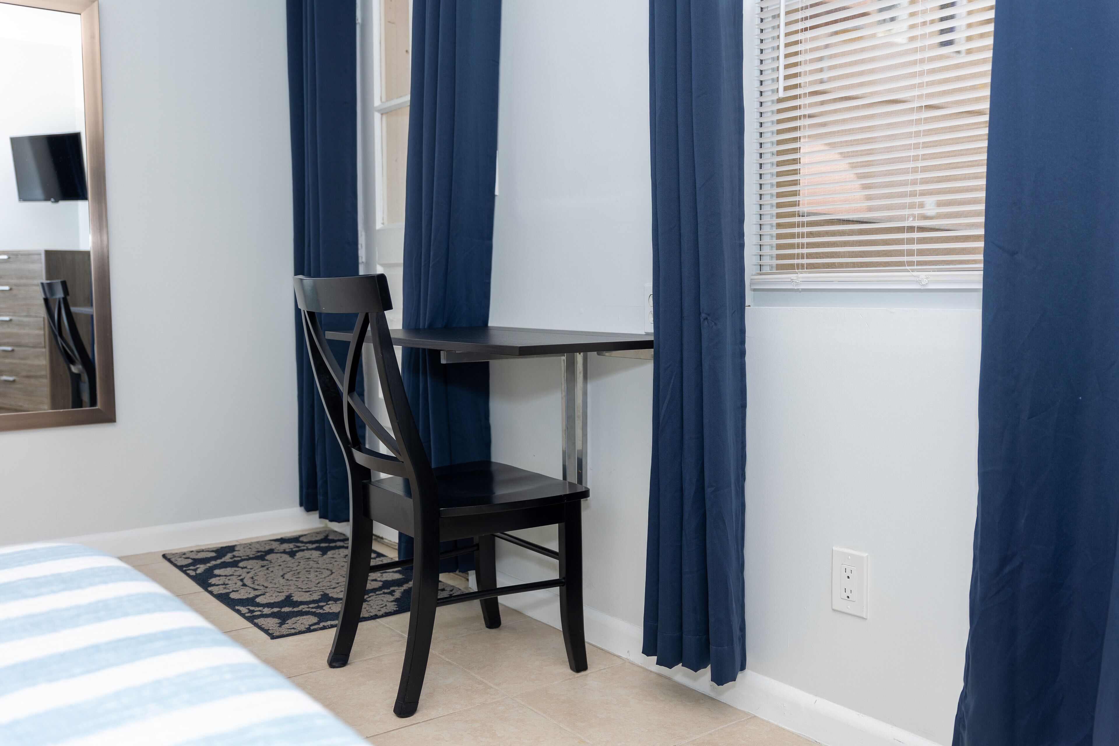 Quarto com cama, escrivaninha, cadeira e janela com cortinas.