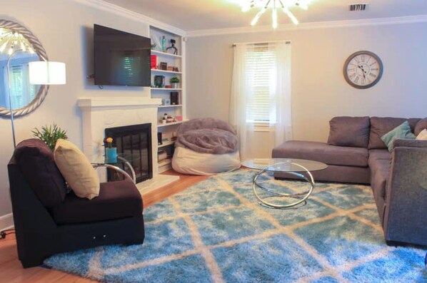 A living room with a fireplace, a television, a sofa, a bean bag, and a clock on the wall. - Beautifully Designed Oasis - Blocks to UF Stadium! (Gainesville)