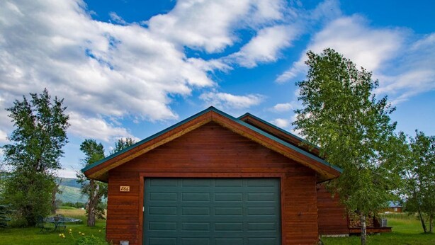 En stuga av trä med en grön garagedörr, omgiven av träd och en klar himmel.