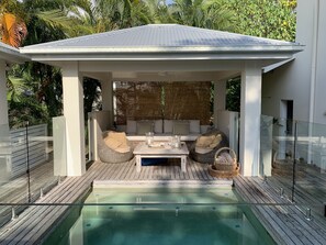 A covered outdoor seating area with a pool, wooden deck, and a white gazebo.