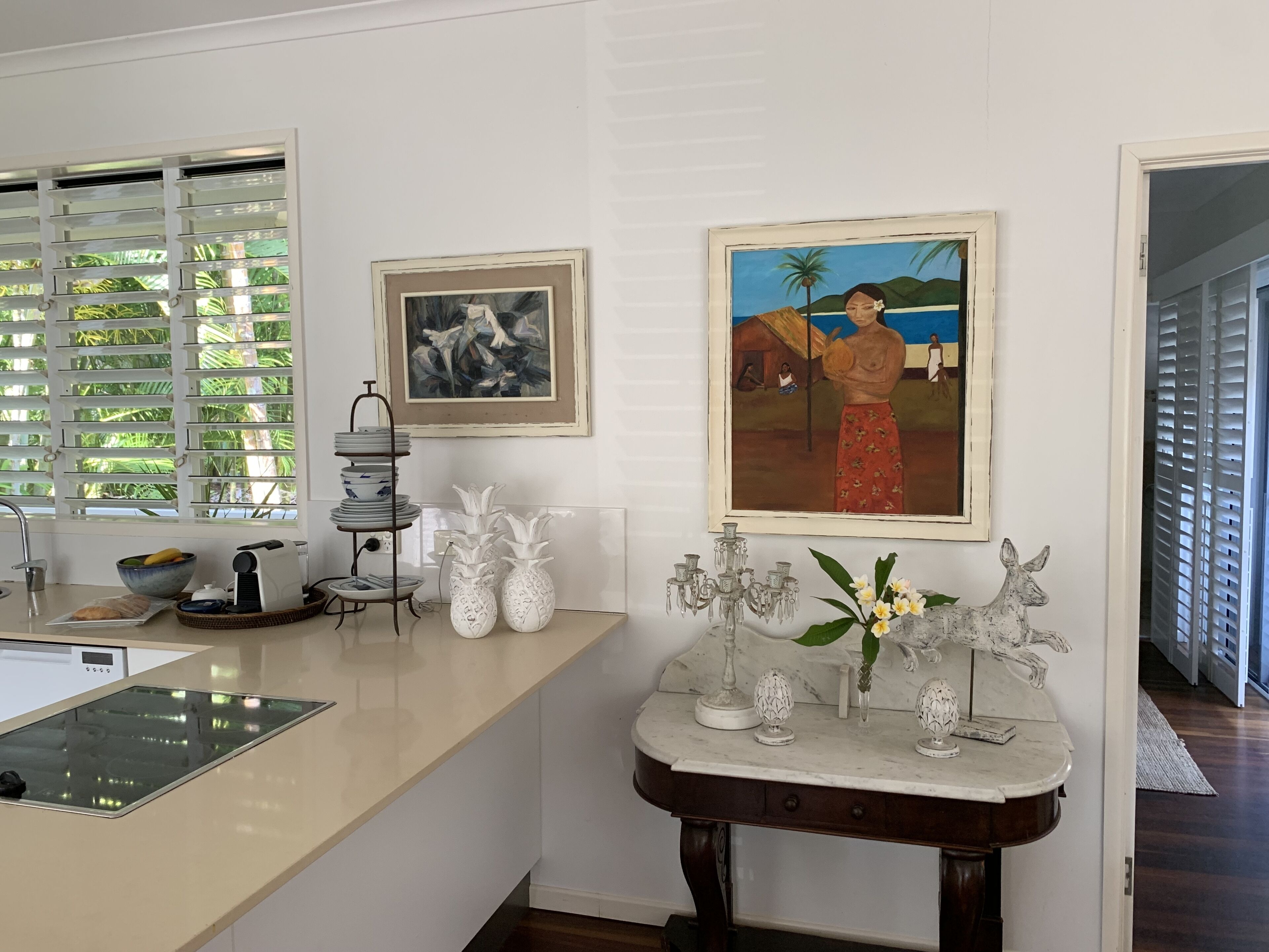 Una cocina con encimera blanca, un lavabo y una ventana con persianas. Hay jarrones decorativos y una lámpara de araña sobre la encimera.