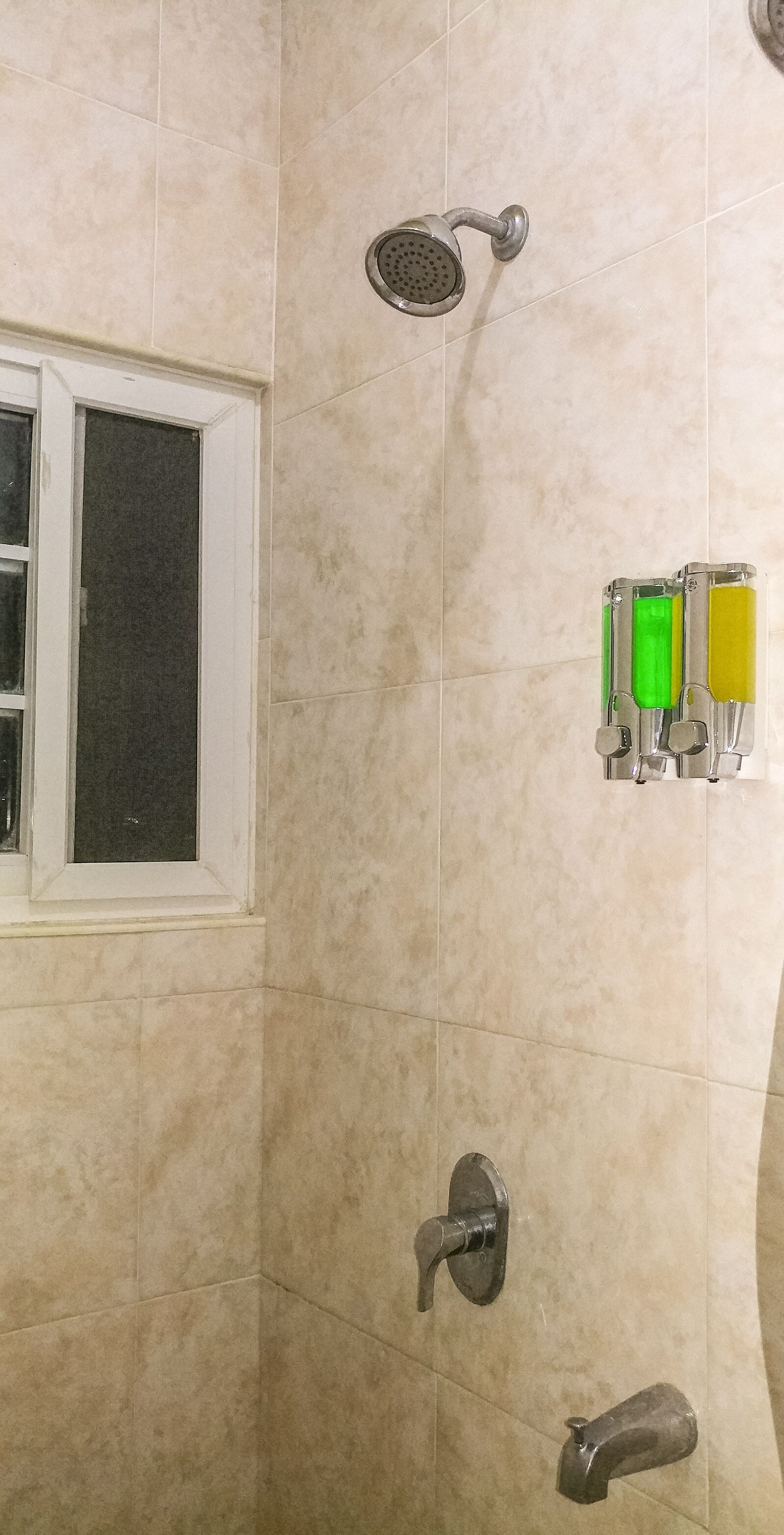 A bathroom with beige tiles, a showerhead, a window, and a set of three dispensers.