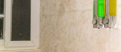 A bathroom with beige tiles, a showerhead, a window, and a set of three dispensers.