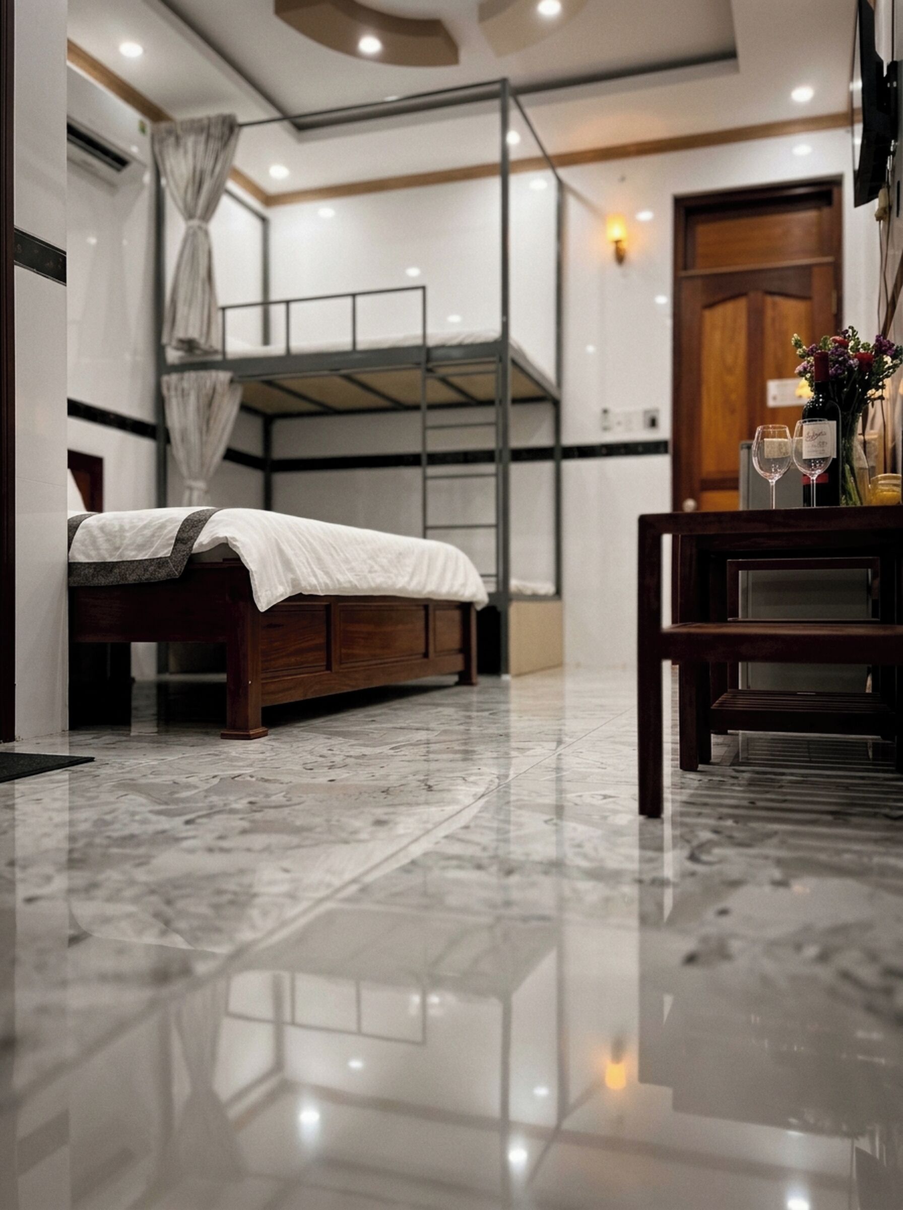A room with a bunk bed, a wooden desk, a chair, a vase with flowers, and a bottle of wine.