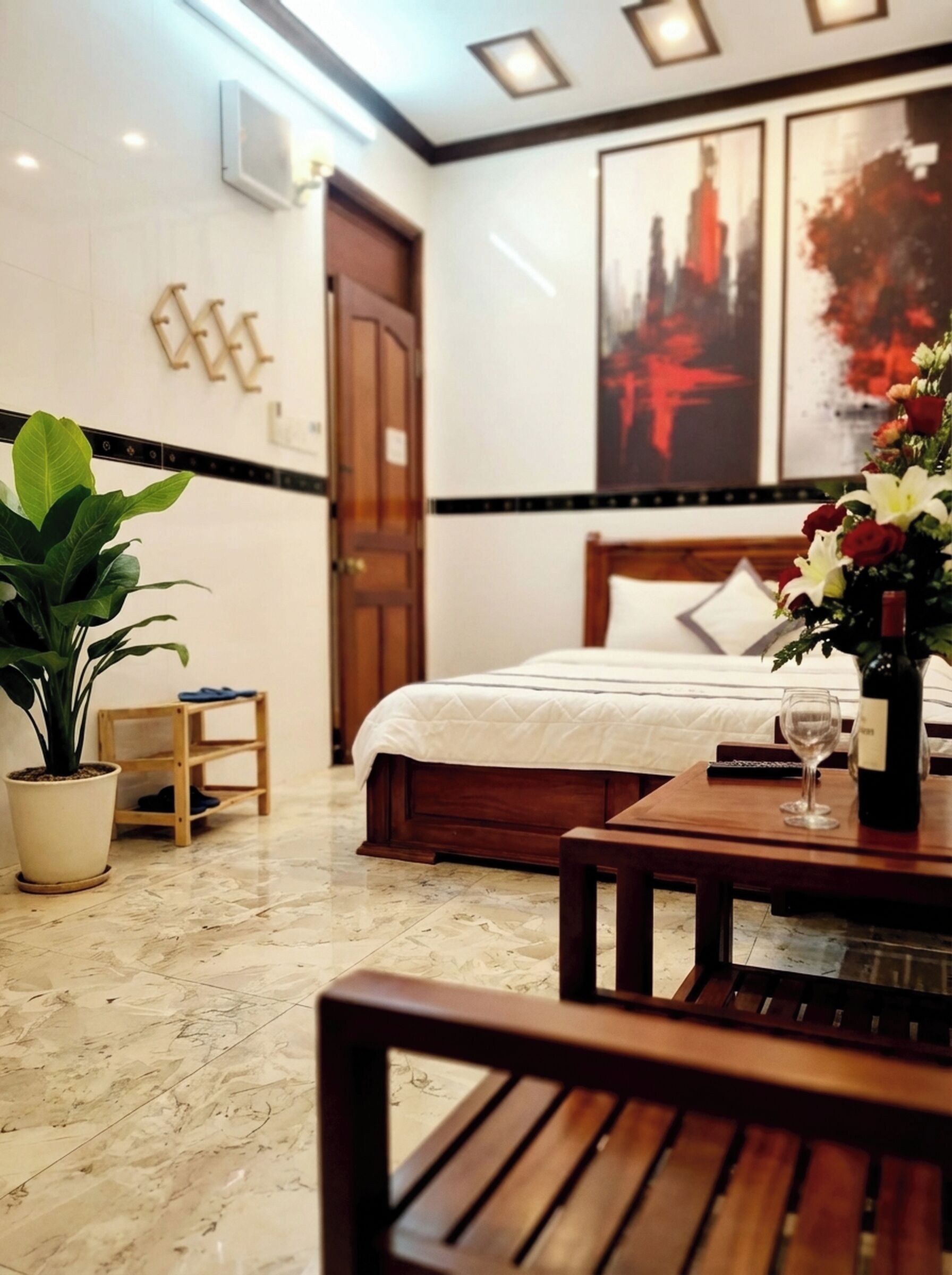 A hotel room with a bed, a wooden nightstand, a potted plant, a small table with a wine bottle and glass, and a large painting on the wall.