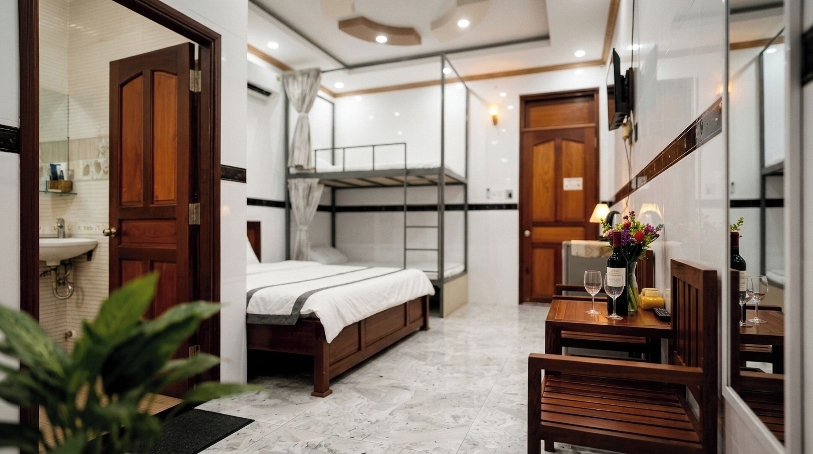 A compact hotel room with a bed, a small table with a bottle and glasses, a mirror, and a wooden door.
