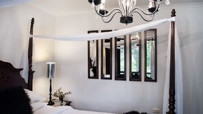 Quarto com cama de dossel, candelabro, espelho e mesa de cabeceira com abajur e planta.