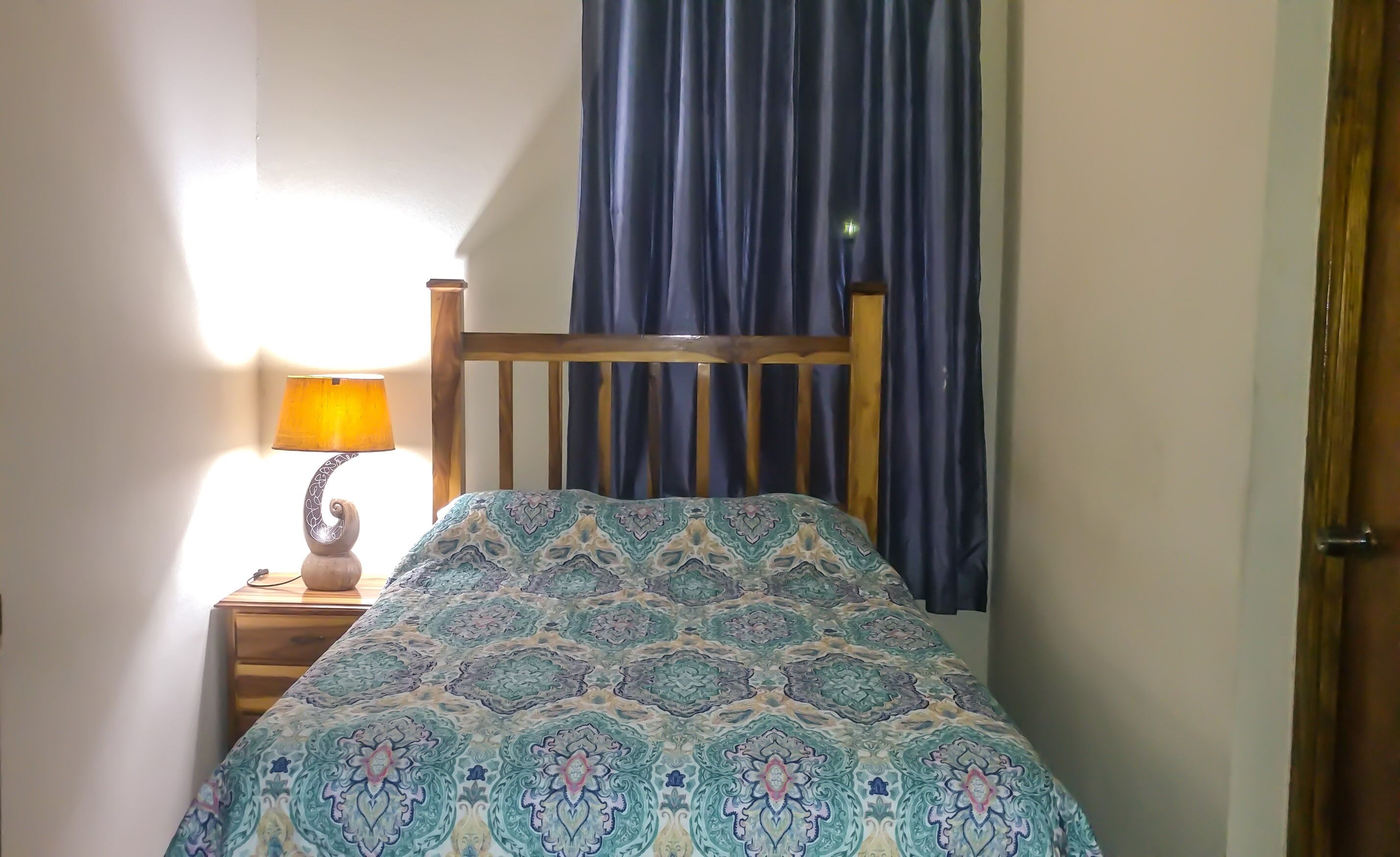 Un dormitorio con cabecera de madera, colcha estampada, lámpara de noche y ventana con cortinas azules.