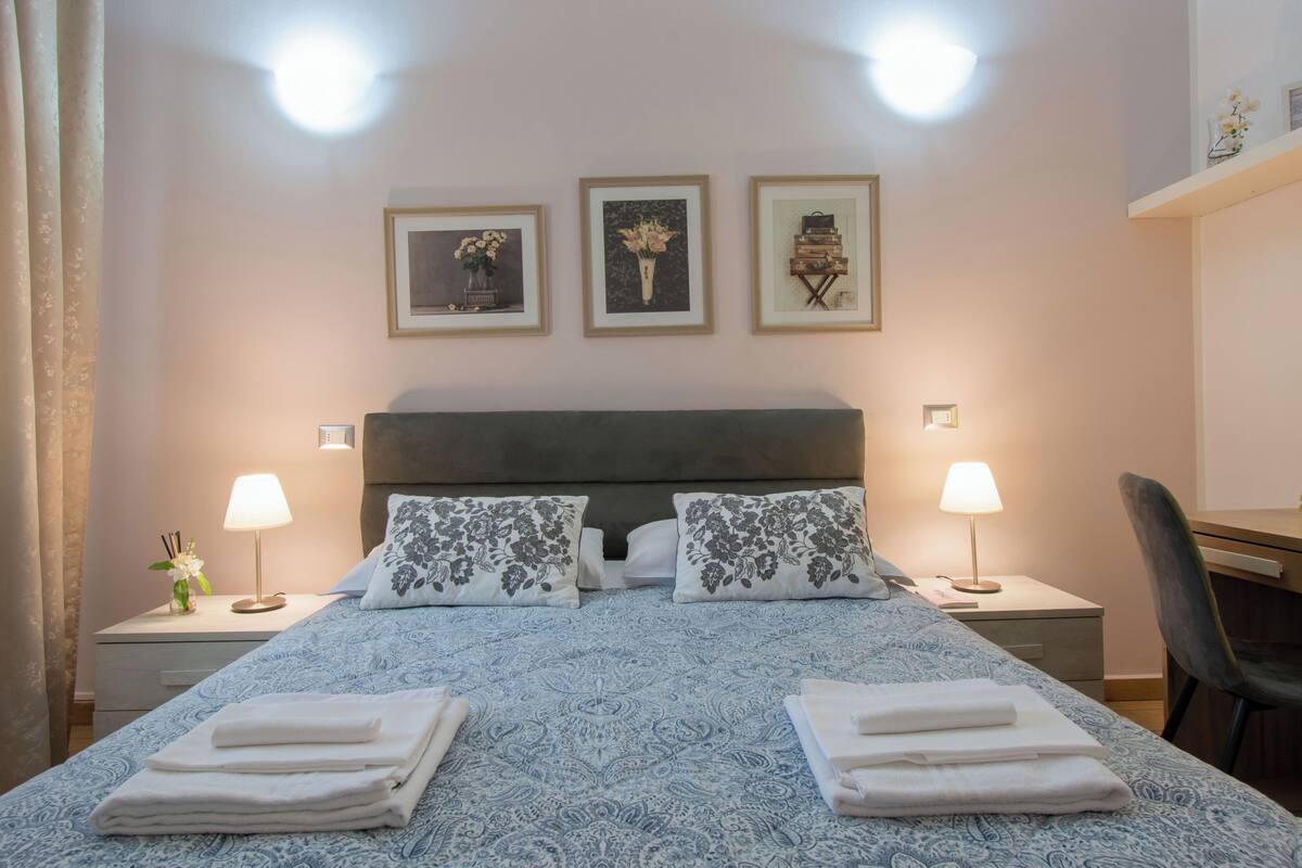 A hotel room with a bed, two nightstands, a desk, and a chair. There are three framed pictures on the wall, a lamp on each nightstand, and a small plant on the left nightstand.