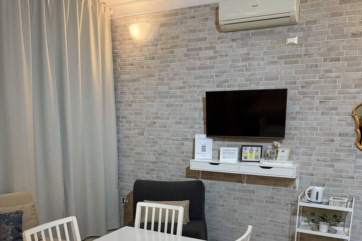 A living room with a white table and chairs, a sofa, a flat-screen TV mounted on a brick wall, and a white shelf with a kettle and a plant.