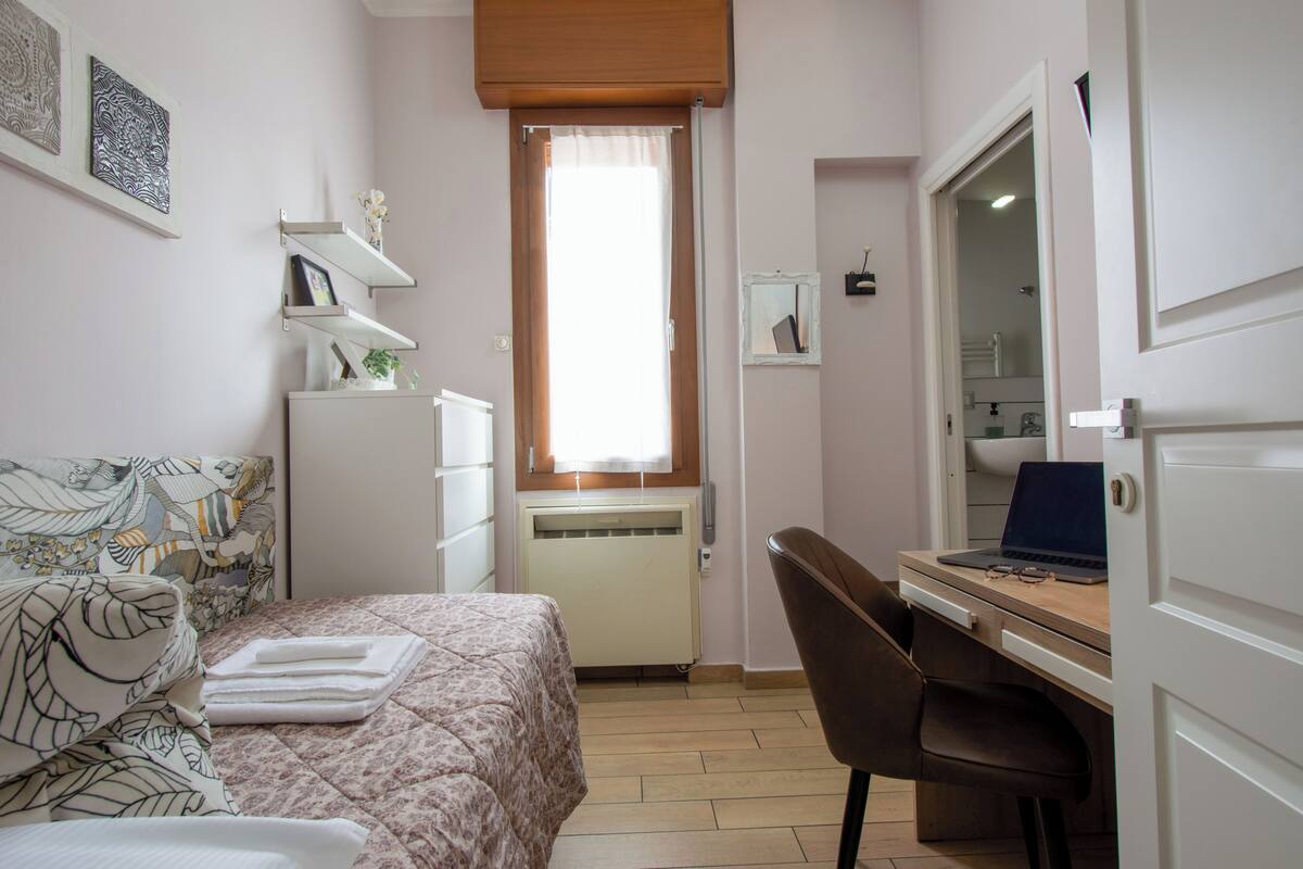 A bedroom with a bed, a desk with a laptop, a chair, a radiator, and a window with curtains.