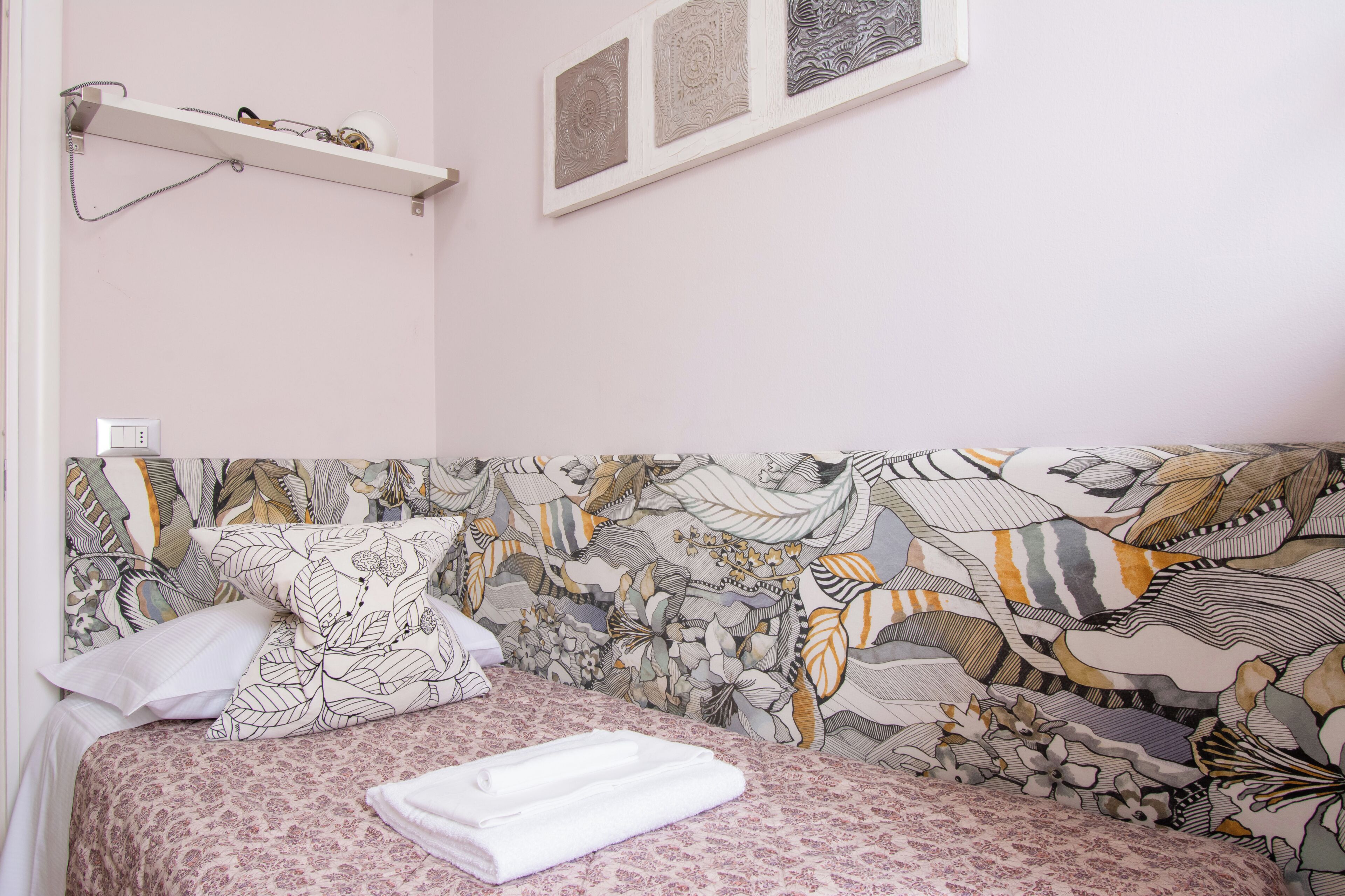 Un dormitorio con cabecera estampada, mesita de noche blanca y estante con lámpara.