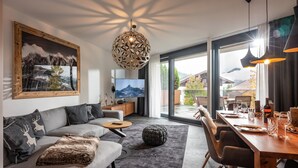 Un salon moderne comprenant un canapé, une table basse et un pouf, un coin repas avec une table et des chaises en bois, un téléviseur et une grande fenêtre offrant une vue sur les montagnes.