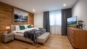 Une chambre moderne avec une tête de lit en bois, un grand lit avec une literie verte et blanche, une table de chevet avec une lampe, un banc, une commode et un téléviseur posé sur un meuble en bois.