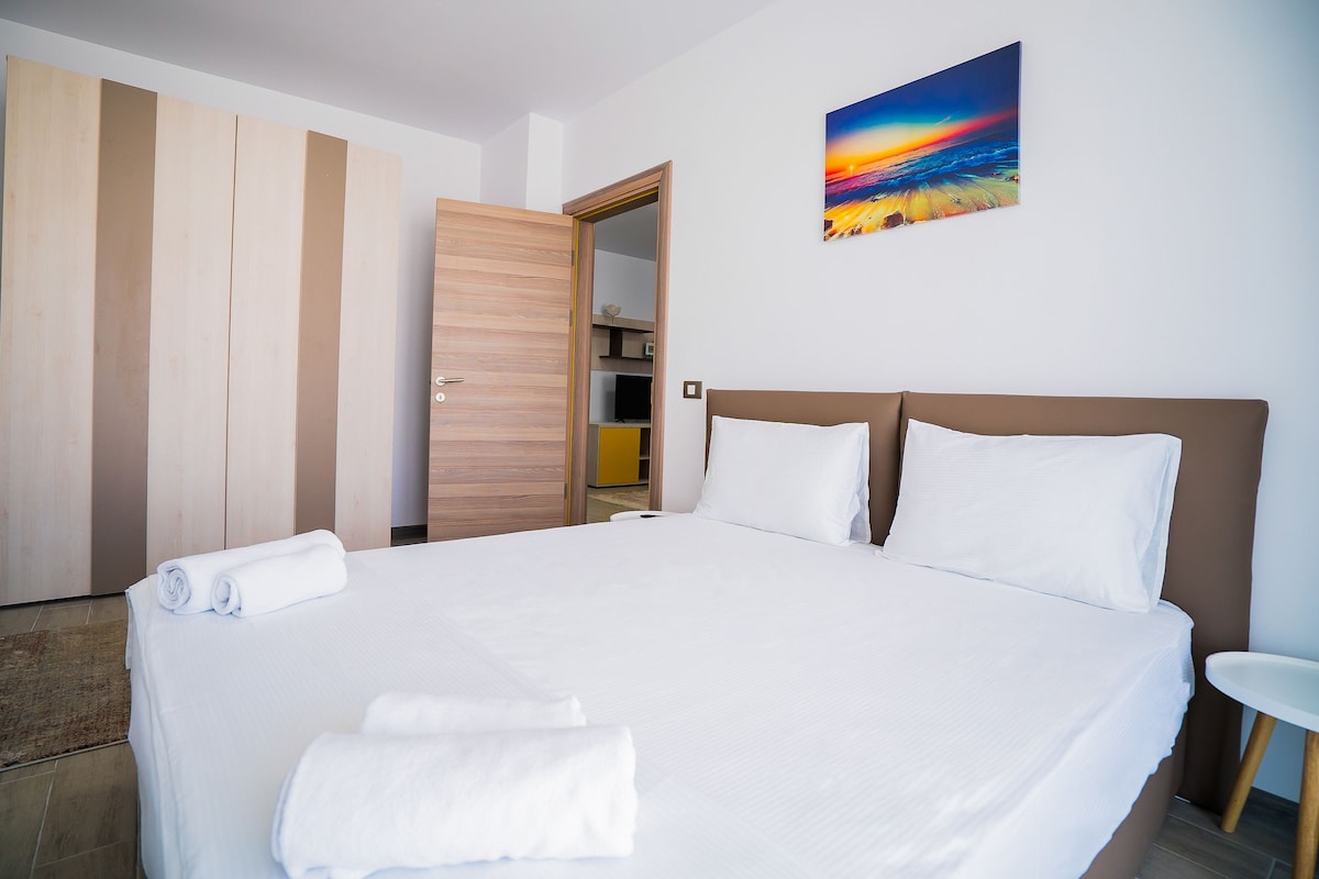 A neatly made bed with white linens, a wooden wardrobe, and a painting of a sunset on the wall.