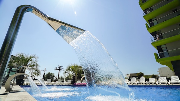 A pool with a water feature, surrounded by lounge chairs and palm trees.
