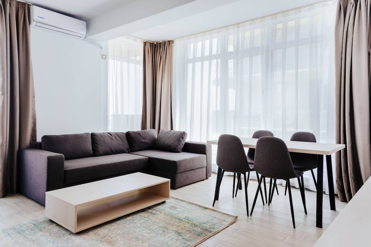 Una sala de estar moderna con un sofá seccional, una mesa de centro, una mesa de comedor con sillas y ventanas amplias con cortinas translúcidas.